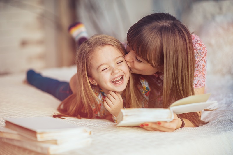 Portrait of a smiling young cute mother and daughter reading a book lying and relax in the bed in a bright big white room